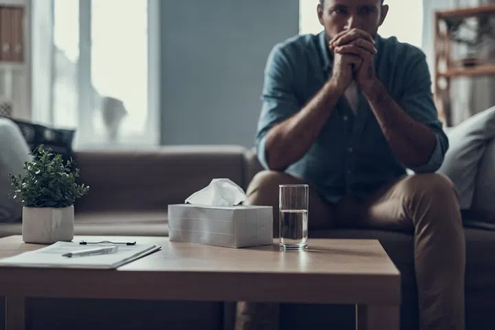 Man sitting on the sofa with his fingers crossed in front of his face and having a glass of water