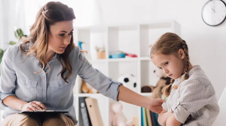 adult psychologist with clipboard talking to sad little child in office
