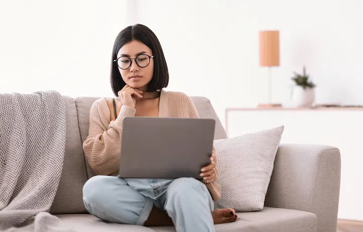 Pensive asian female freelancer in glasses with laptop brainstorming at new project, making research in internet, having remote job, sitting on couch in living room at home, looking at computer screen