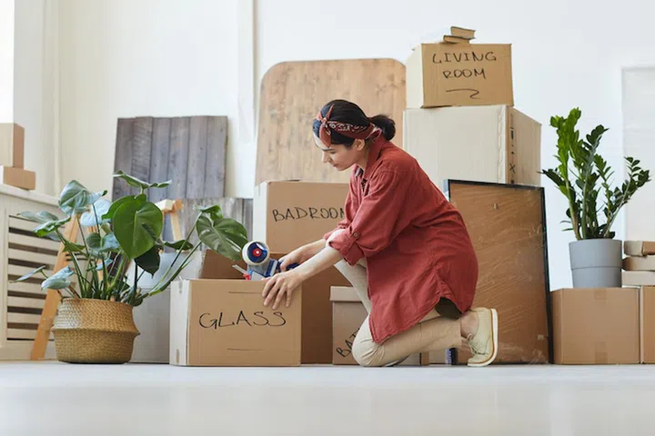 Young woman sealing the boxes with things with adhesive tape during relocation