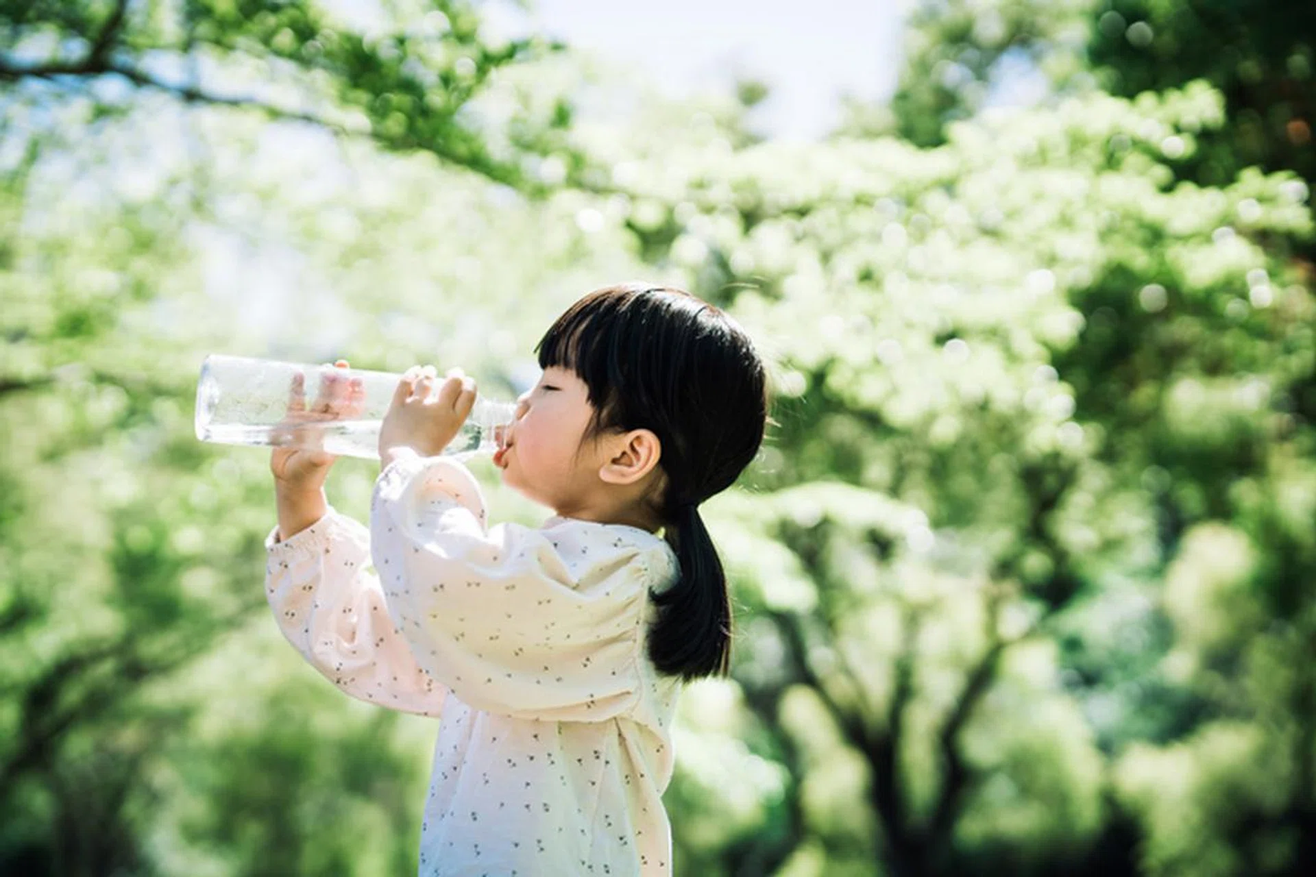Side profile of little Asian girl drinking water from a reusable plastic bottle in the park outdoors on a sunny day. Zero waste and sustainable lifestyle concept
