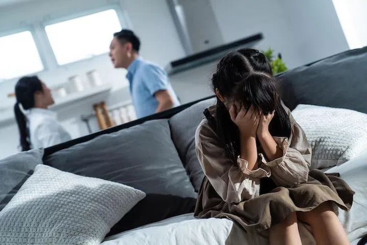 Asian girl kid sitting and crying on sofa while parents having fighting or quarrel conflict at home. Child covering face and eyes with hands do not want to see the violence. Domestic problem in family