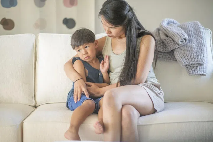 A middle aged Asian mother sits with her four year old son on their living room couch while she has a serious conversation with him.