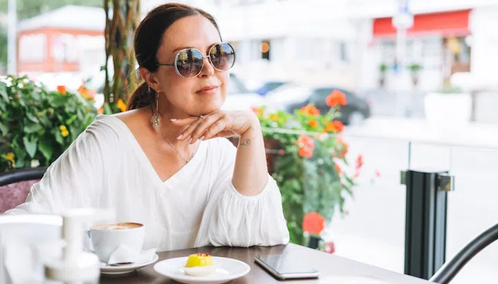 Portrait of smiling beautiful brunette middle aged woman in sunglasses in white clothes with coffee at terrace of summer cafe