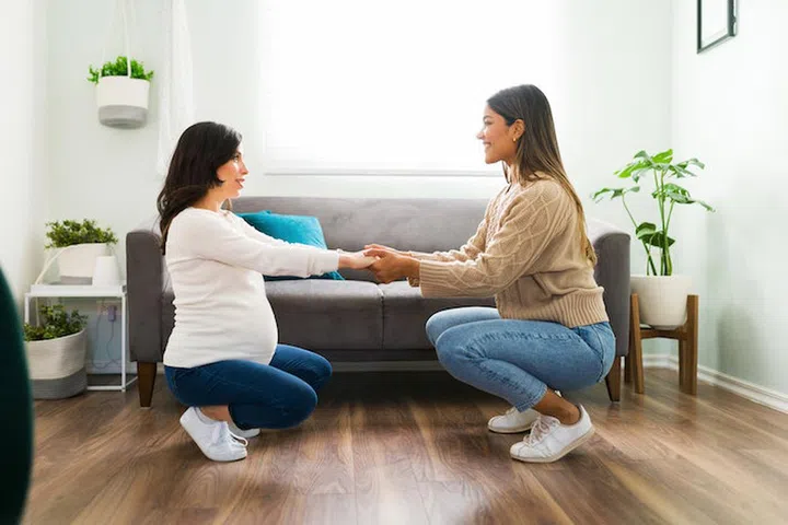 Side view of a happy doula smiling and holding hands with a caucasian pregnant woman while doing stretching exercises in the living room