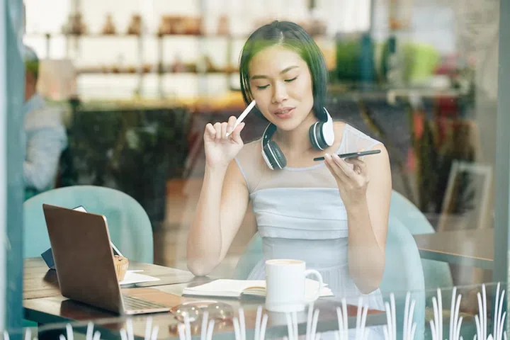 Creative pretty young student sitting at cafe table and recording her ideas and thoughts via smartphone