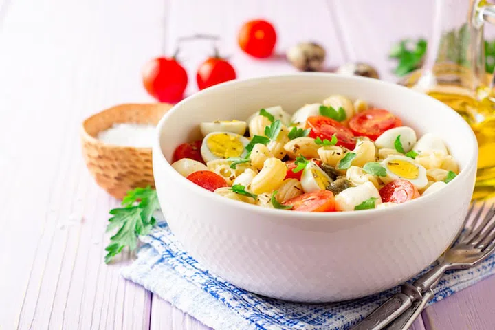 Pasta salad with quail eggs, mozzarella, cherry tomatoes and capers in bowl on purple wooden background. Selective focus.