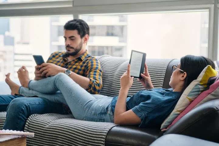 Young couple holding ereader, reading ebook while lying barefoot on sofa at home