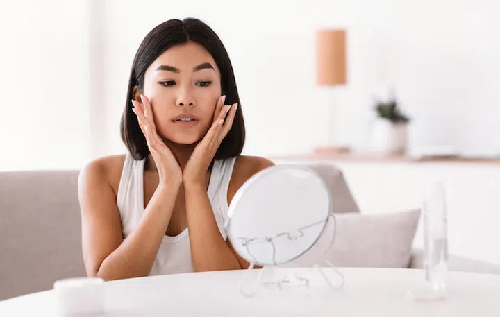 Facelift And Skin Care Concept. Portrait Of Young Asian Lady Massaging Her Face Sitting In Front Of The Mirror. Lady Applying Moisturizing Cream, Doing Facial Anti Aging Massage At Home