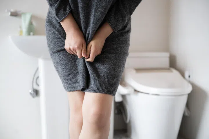A woman wearing knitwear is complaining of pain from urinary incontinence in front of the toilet