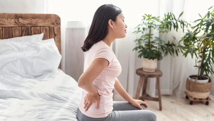 Backache Concept. Asian Lady Suffering From Pain In Lower Back Sitting On Bed At Home.