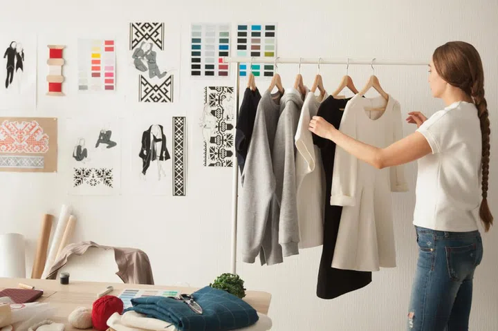 Female fashion designer works on new womenswear collection for clients in cozy workshop studio, dressmaker, tailor or needlewoman standing near clothing rack with fashionable stylish handmade clothes