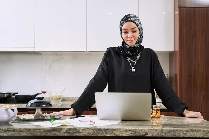 Cheerful young woman in hijab is working online in kitchen and using notebook and smartphone