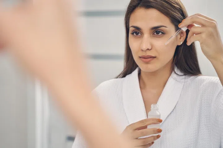 Attractive lady looking in the mirror and having skincare procedure while holding glass dropper and bottle of clear liquid (Attractive lady looking in the mirror and having skincare procedure while holding glass dropper and bottle of clear liquid, ASC