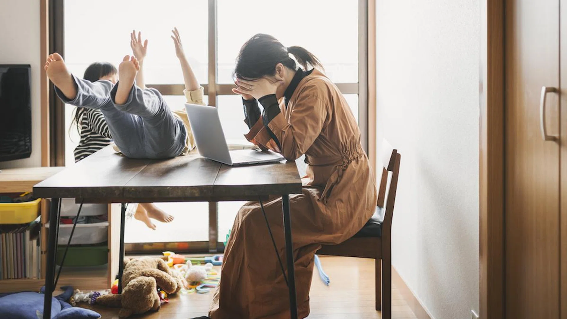 Asian mother working with laptop while her son and daughter are playing in the living room. Her son is playing on the table.