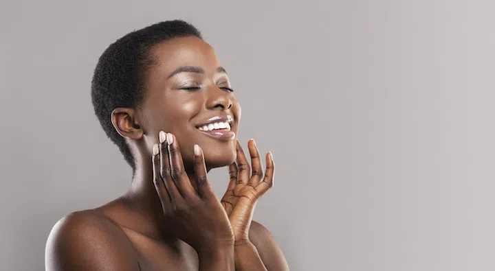Face lifting concept. Happy black woman with closed eyes touching soft smooth skin on her cheeks over grey background, panorama