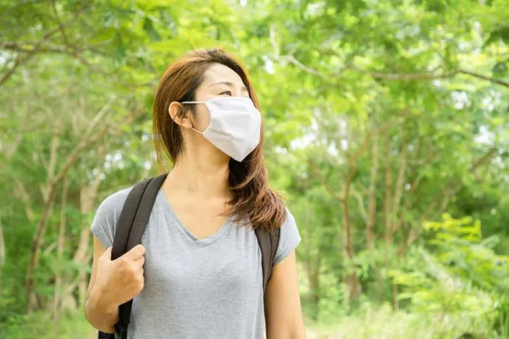 Asian woman tourism traveling in a beautiful green park wearing protective mask with backpack walking alone enjoying the nature, new normal lifestyle concept