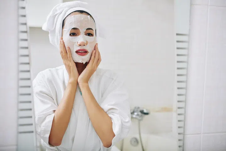 Young Asian woman with sheet mask on her face looking in mirror