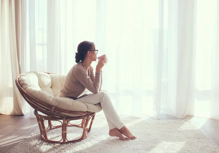 Young woman at home sitting on modern chair in front of window relaxing in her lliving room and drinking coffee or tea