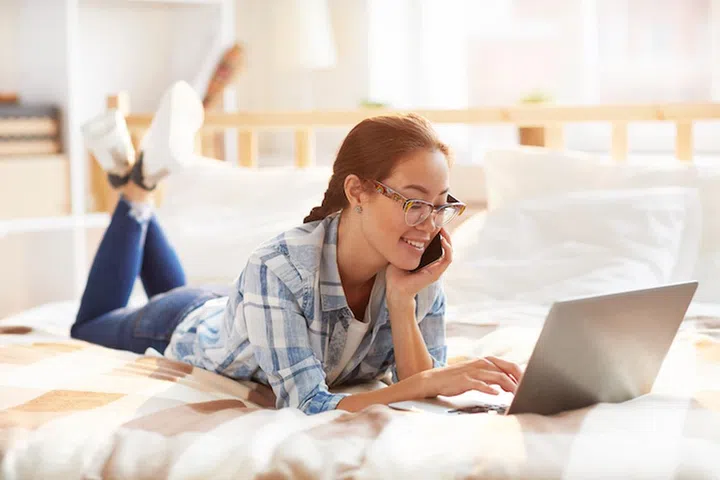 Full length portrait of contemporary Asian woman using laptop lying on  bed  in sunlight and relaxing, copy space