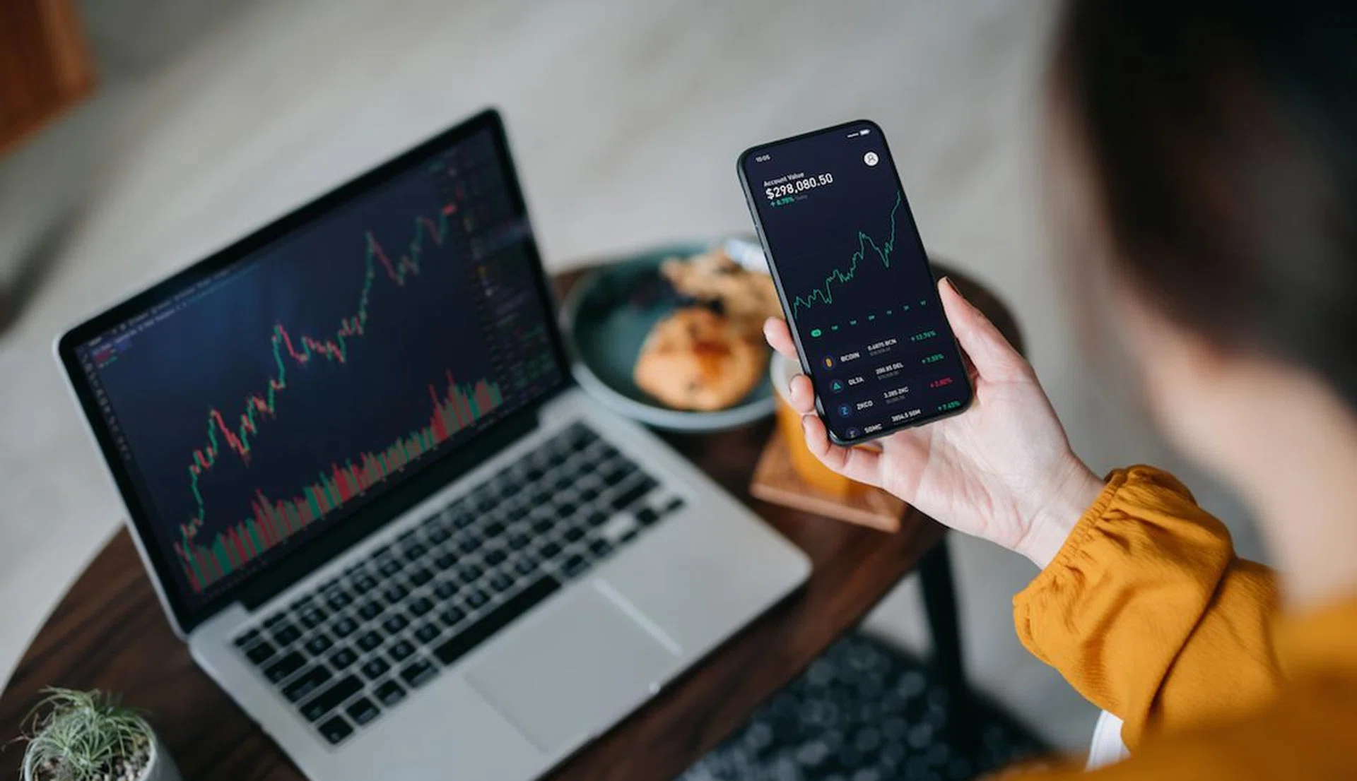 Over the shoulder view of Asian woman holding smartphone, analyzing investment trading data on crypto graph with smartphone and laptop while working at home. Cryptocurrency, bitcoin, digital ledger