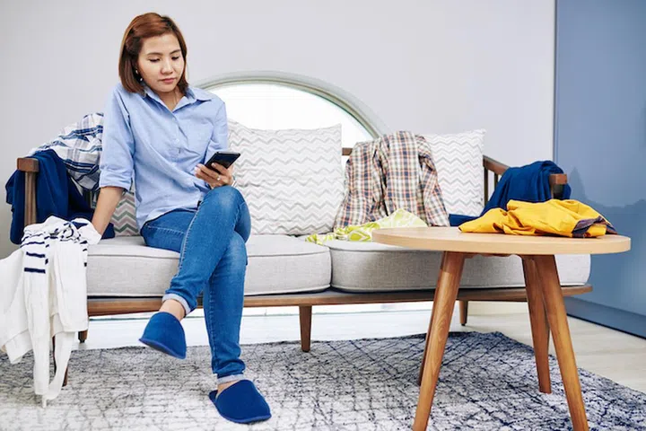 Lazy young Vietnamese woman sitting on sofa with her clothes around and reading posts on social media