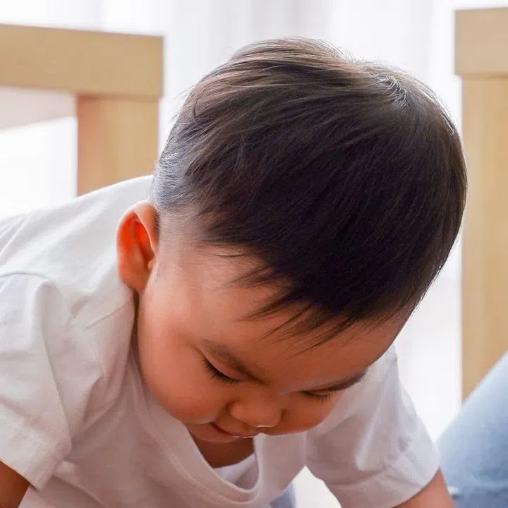 Serious Asian little boy crawling on floor while young father sitting next to him in living room at home