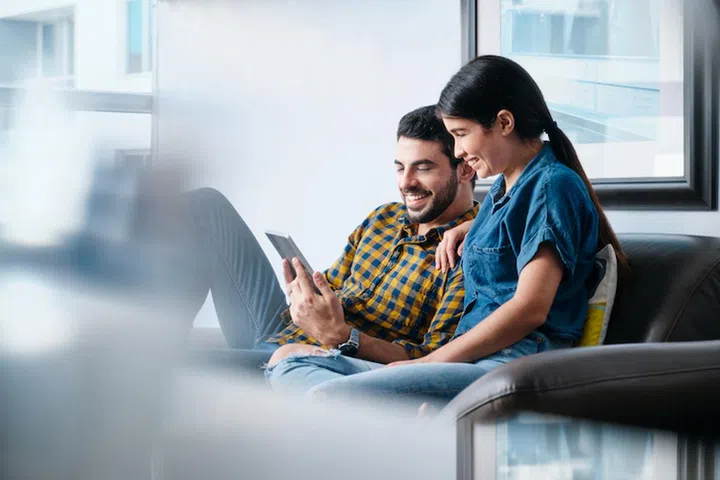 Young couple sitting on couch, smiling while streaming a movie from the Internet on Tablet PC. Copy space.