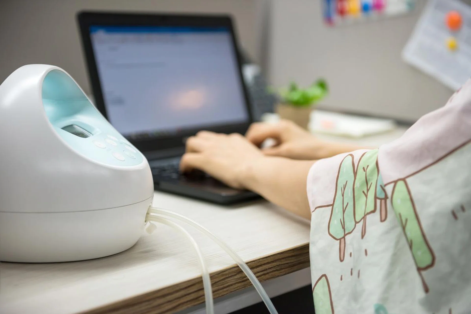 Busy mother or mom pumping breastmilk by Automatic breast pump machine wirh Nursing fabric cover while typing on laptop pc computer on table. Motherhood in corporate office.