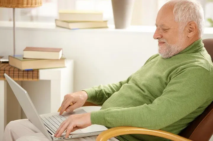 8748737 - smiling pensioner using laptop computer, sitting in armchair at home.
