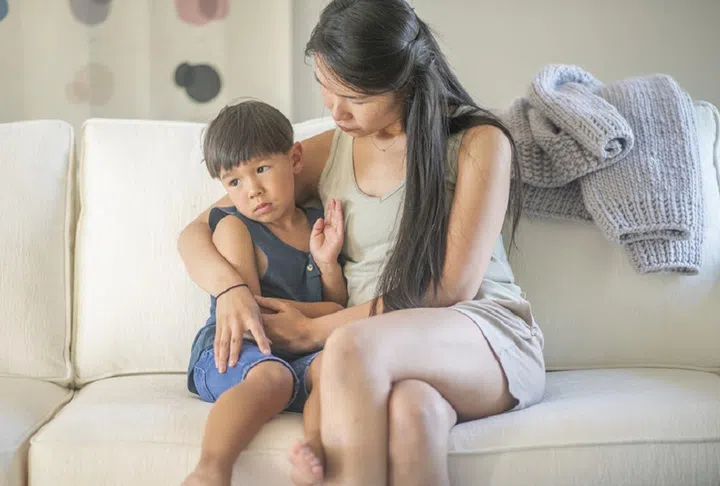 A middle aged Asian mother sits with her four year old son on their living room couch while she has a serious conversation with him.
