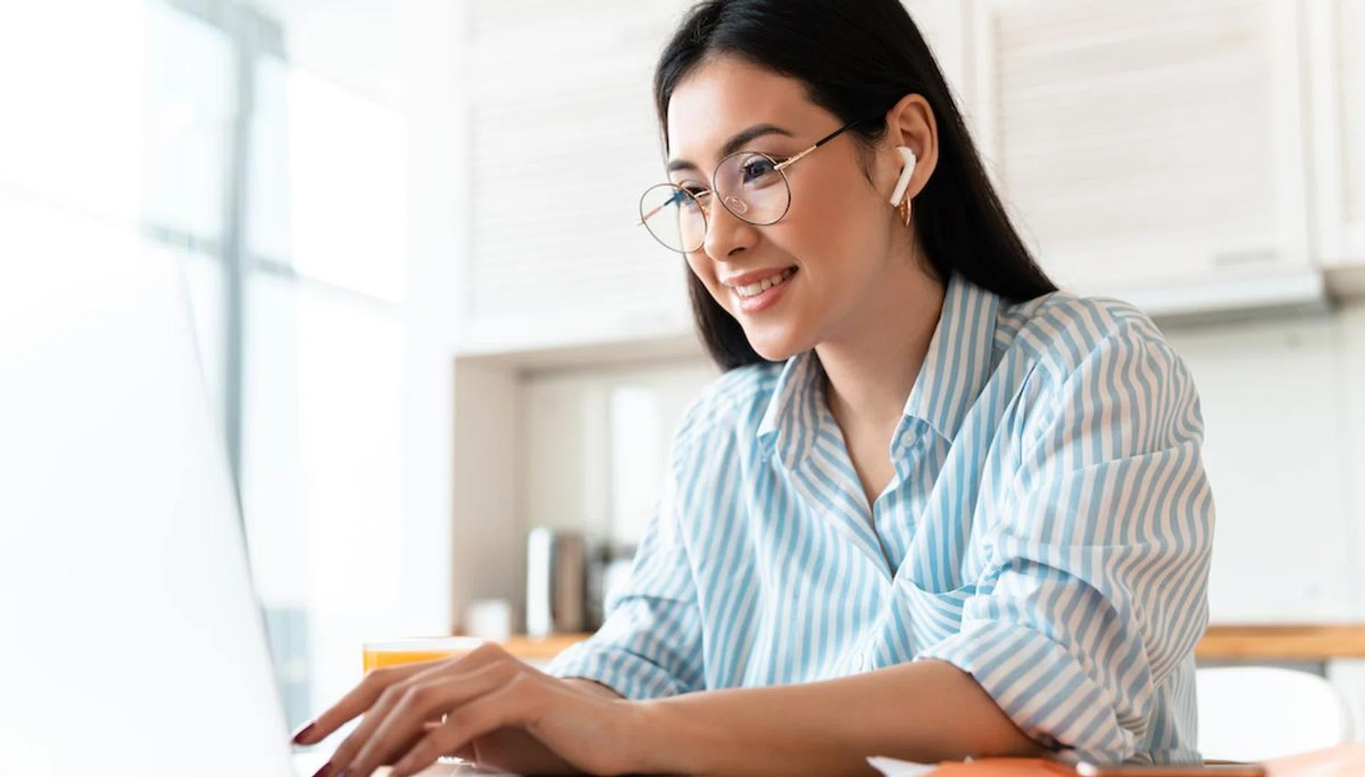 Image of a beautiful happy brunette young woman at the kitchen indoors at home using laptop computer and wireless earphones.