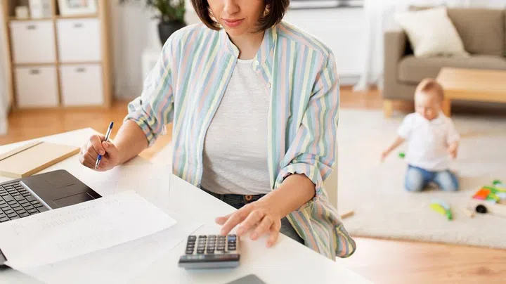 multi-tasking, freelance and motherhood concept - working mother with laptop computer counting on calculator and baby boy playing at home office