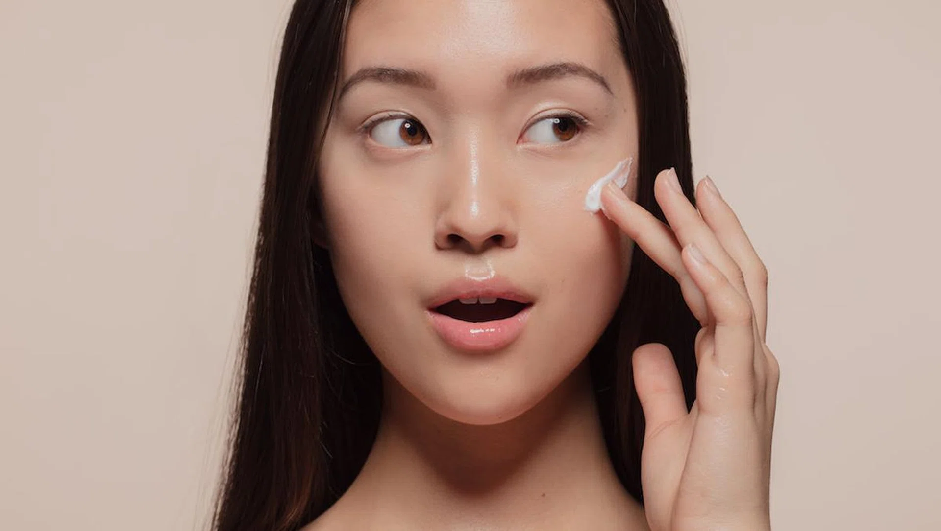 Close up of a asian female model applying moisturizer to her face and looking away. Woman applying moisturizer cream on her pretty face against beige