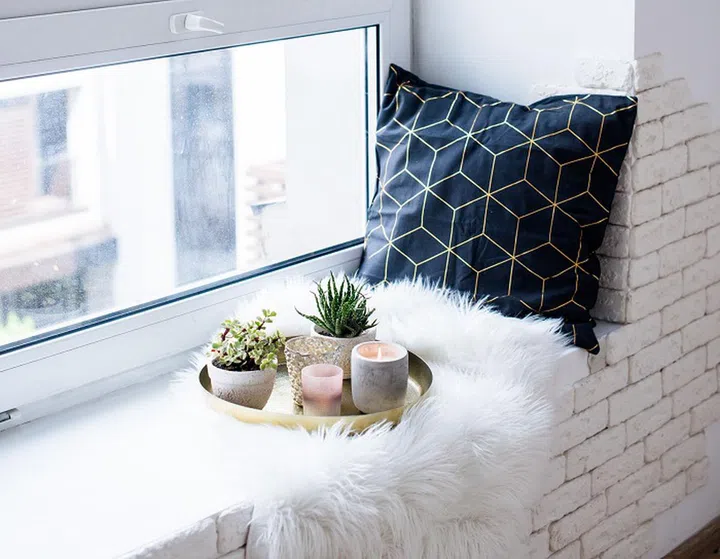 Cozy real home decoration, burning candles on golden tray with pillow on white faux fur on windowsill, apatment interior decoration