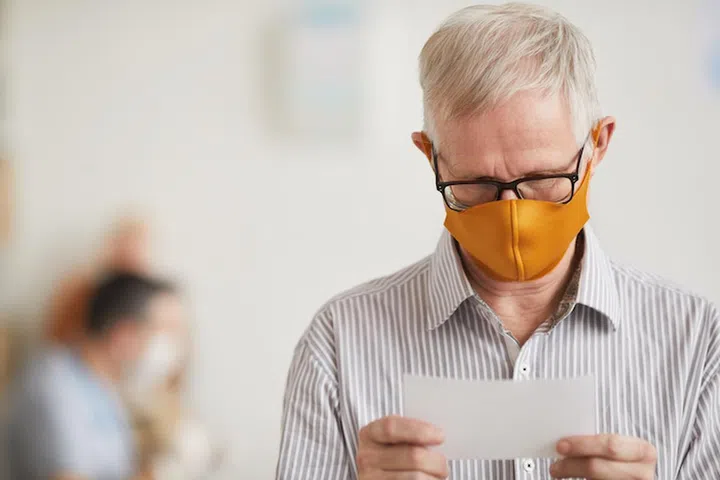 Portrait of white haired senior man wearing mask and reading prescription note at doctors office in medical clinic