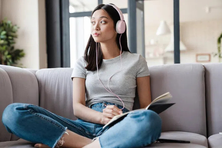 Image of thinking asian student girl in headphones writing down notes while sitting on sofa at home