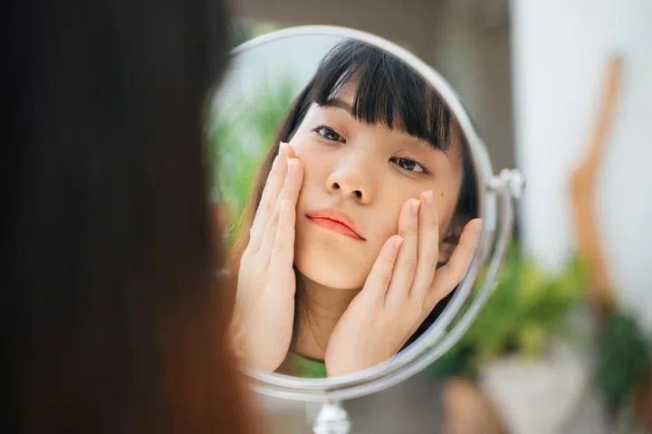 Close up young asian woman applying face cream looking in the mirror.