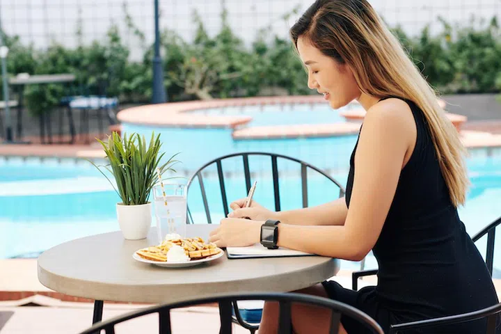 Young Asian woman sitting at table by swimming pool, eating breakfast and filling gratitude journal