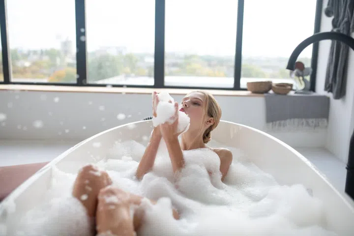 Having much fun. Blonde-haired young woman chilling in foamy bath having much fun