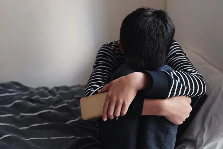 Young Asian preteen teenager boy hugging his knee in his bedroom with smartphone, Cyber bullying in kid, depressed child mental health