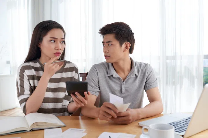 Attractive young woman and her handsome husband looking at each other while calculating family expenses, interior of spacious living room on background