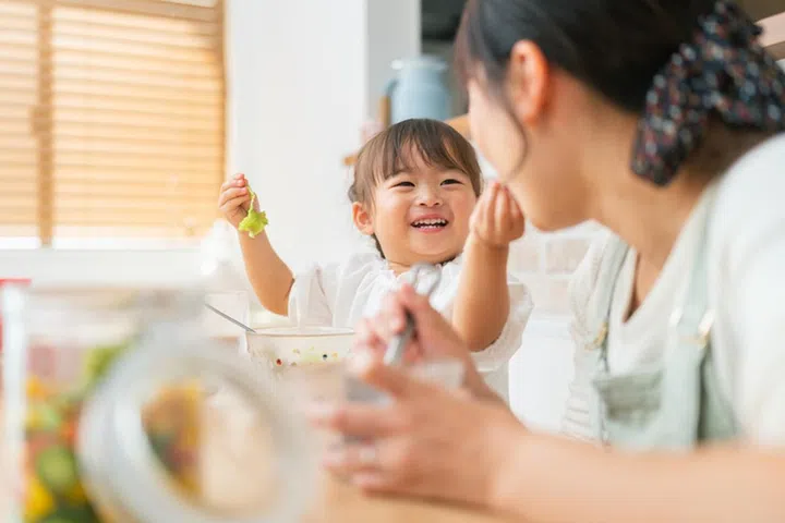 A mother and her small daughter are enjoying eating salad and drinking smoothie at home. A mother is trying to encourage her small daughter for healthy eating.
