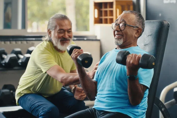 Dumbbells, support and old men in gym, exercise and lifting weights with equipment for strength. Health, smile and senior people in wellness center, fitness or retirement with workout and energy