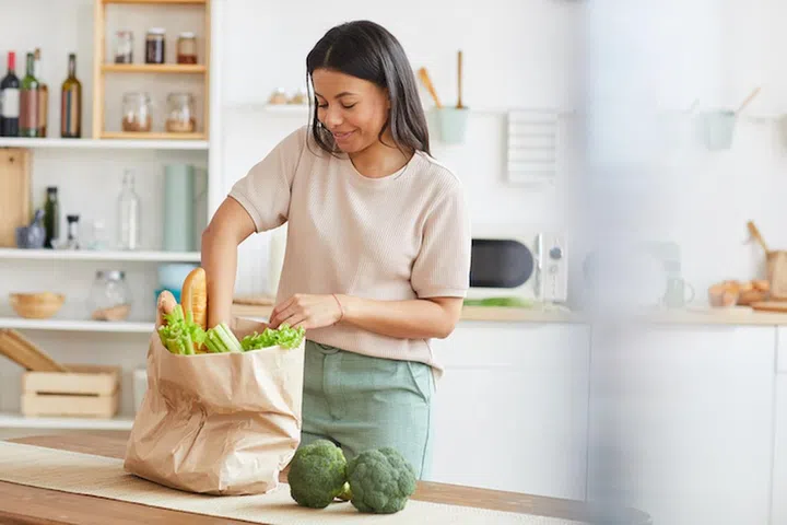 Waist up portrait of elegant mixed-race woman unpacking bags with food while standing in white kitchen interior and smiling, food delivery service, copy space