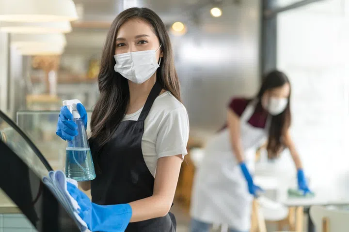attractive positive young Asian woman staff in uniform apron spraying detergent or alcohol on counter product cabinet  wiping it with soft cloth when disinfecting cafe in ther morning safe and clean