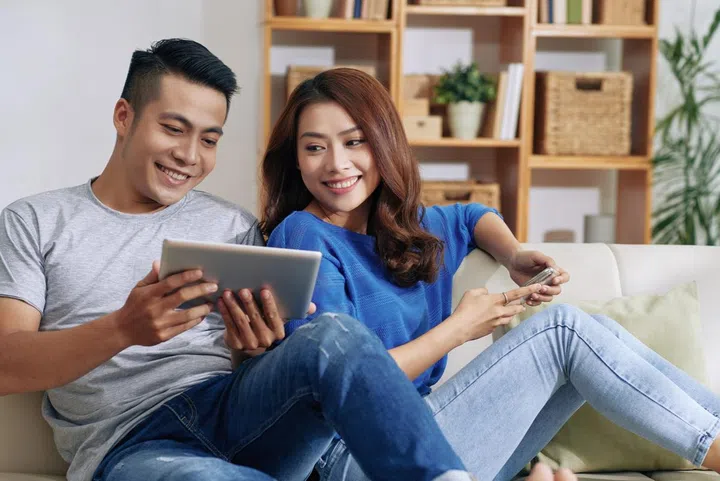 Smiling young man sitting on sofa and showing video on tablet to Asian girlfriend in living room