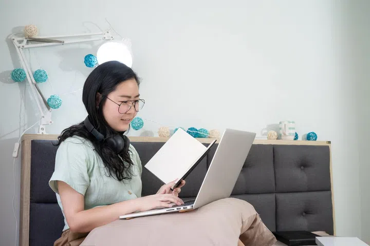 Glasses beautiful Asian woman is working and holding notebook in her hand in the bedroom with a pastel green - brown color theme.