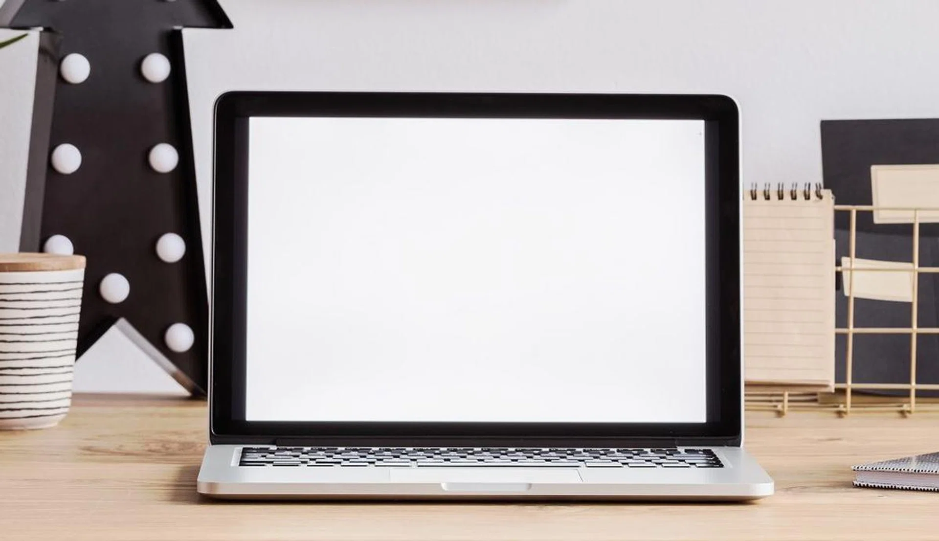 Laptop with white screen on a wooden desk with clock, mug and pencils as well as wall decorations
