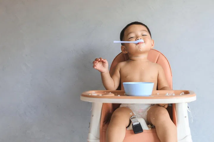Happy infant Asian baby boy eating food by himself on baby high chair and making mess with copy space.